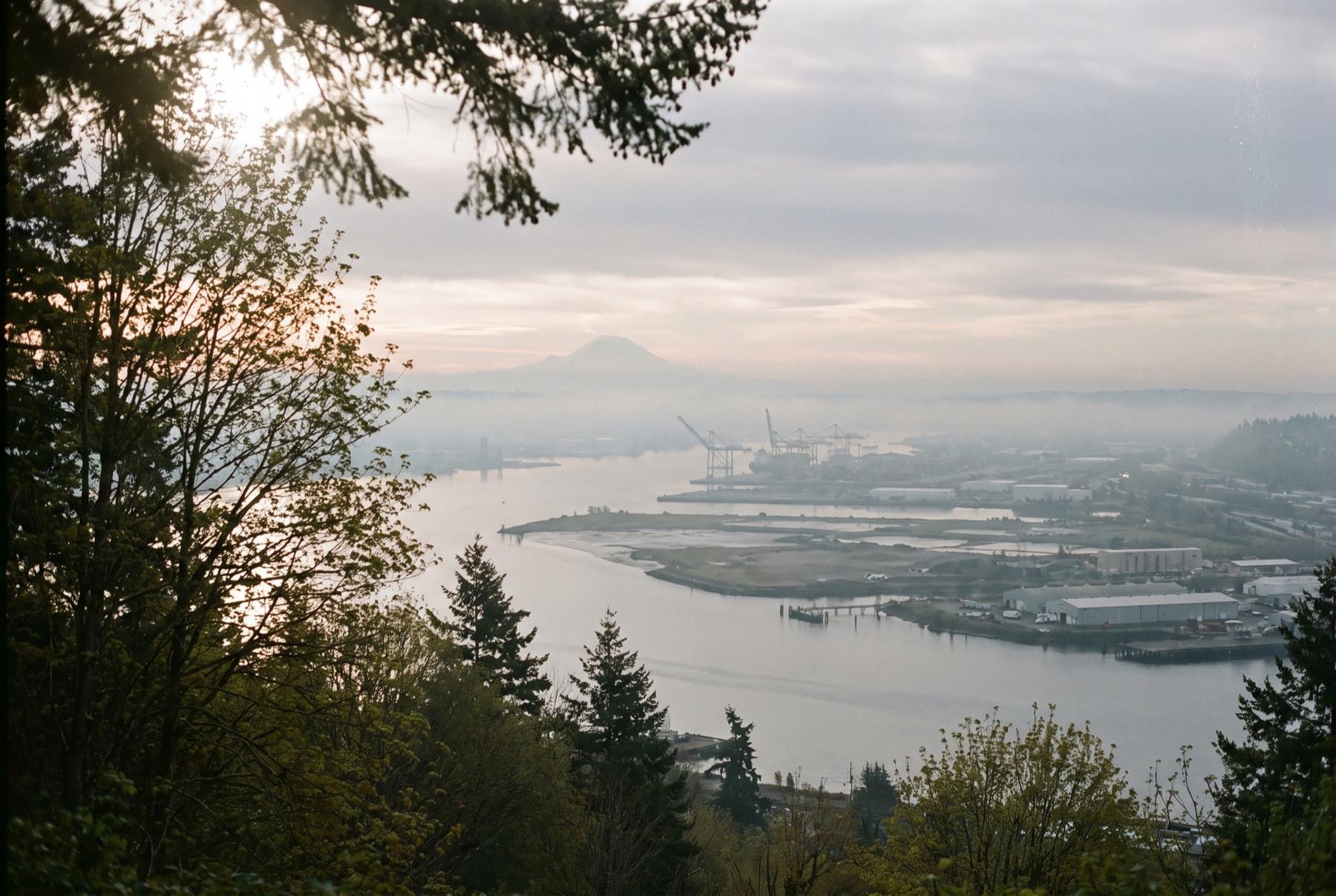 Hazy dawn over Commencement Bay and the Port of Tacoma tideflats, with cranes along the industrial waterways and Mount Rainier faint beyond