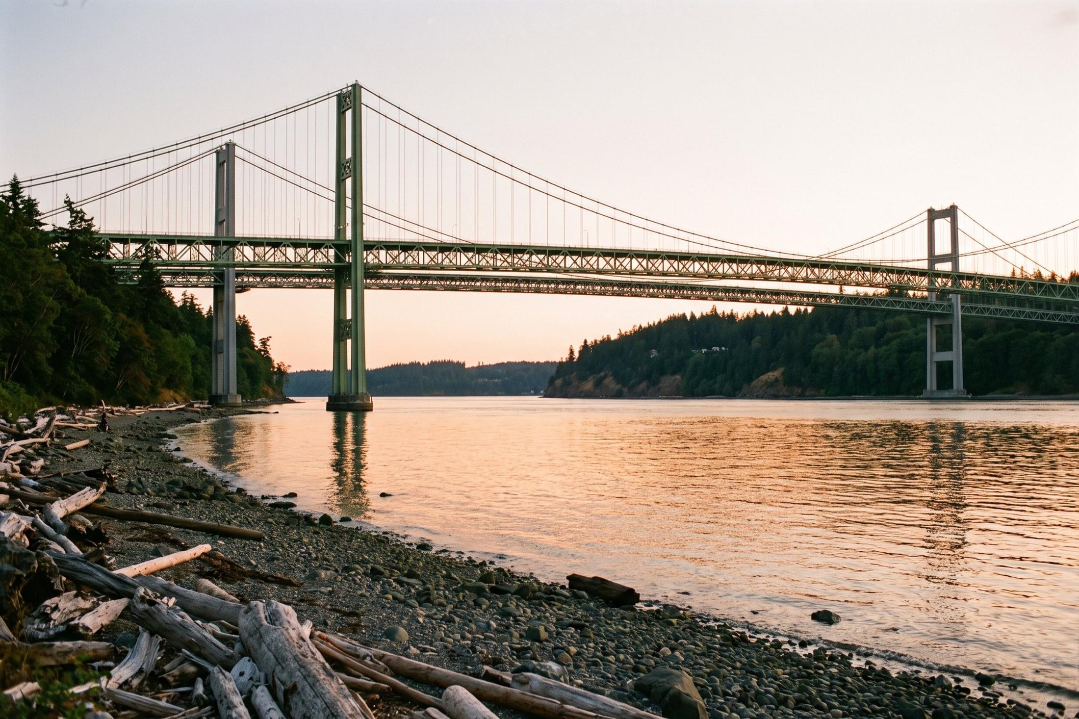 The twin Tacoma Narrows suspension bridges at sunset, seen from a rocky, driftwood-strewn beach below